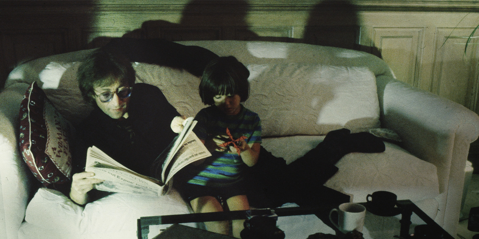 An adult wearing glasses reads a newspaper while sitting on a white sofa next to a child holding a toy. The room has wooden panelling and the lighting casts their shadows on the wall behind them.