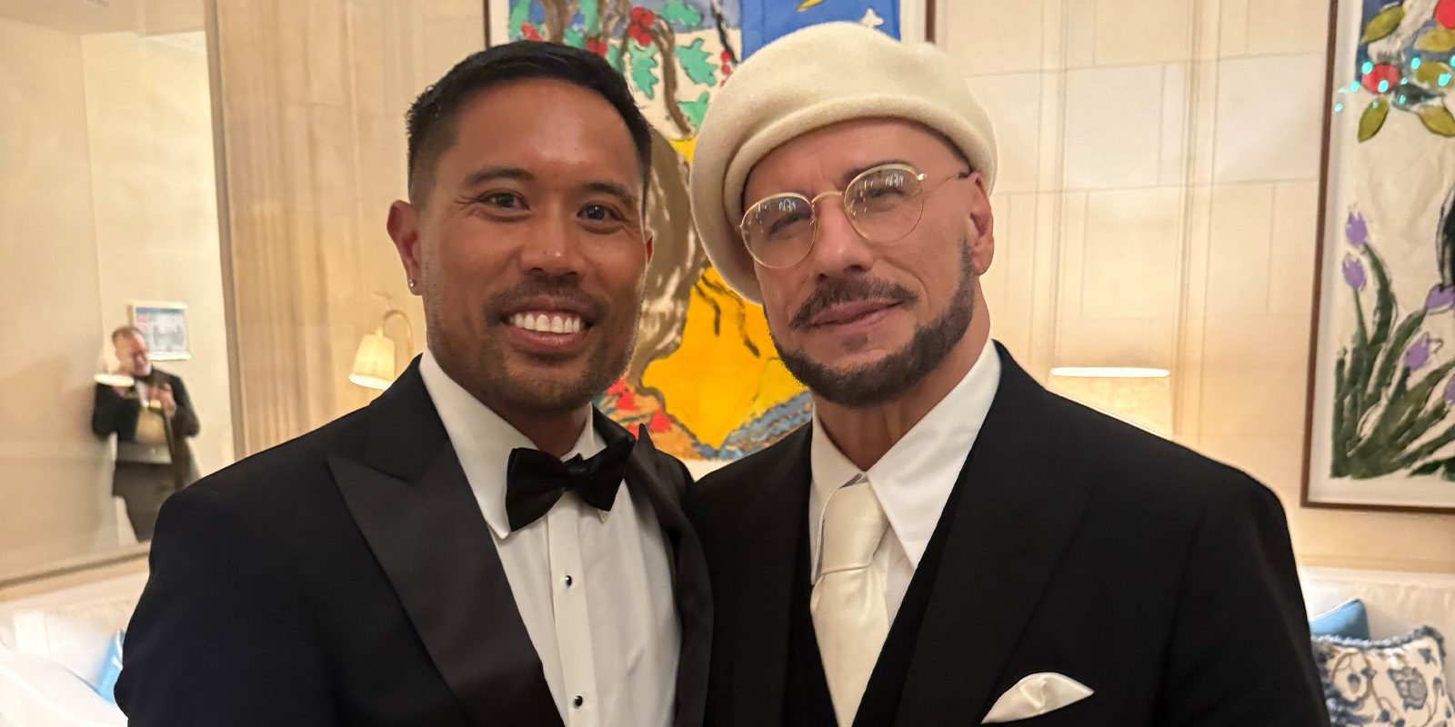 Two men dressed formally in suits and ties stand together, smiling for the camera indoors. One wears a beret and glasses. Colourful artwork hangs on the wall behind them, and a woman in the background takes a photo.