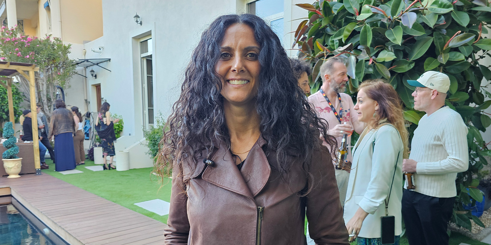 A woman with long curly hair wearing a brown jacket stands outdoors at a social gathering. People are talking and drinking in the background near a building and plants.
