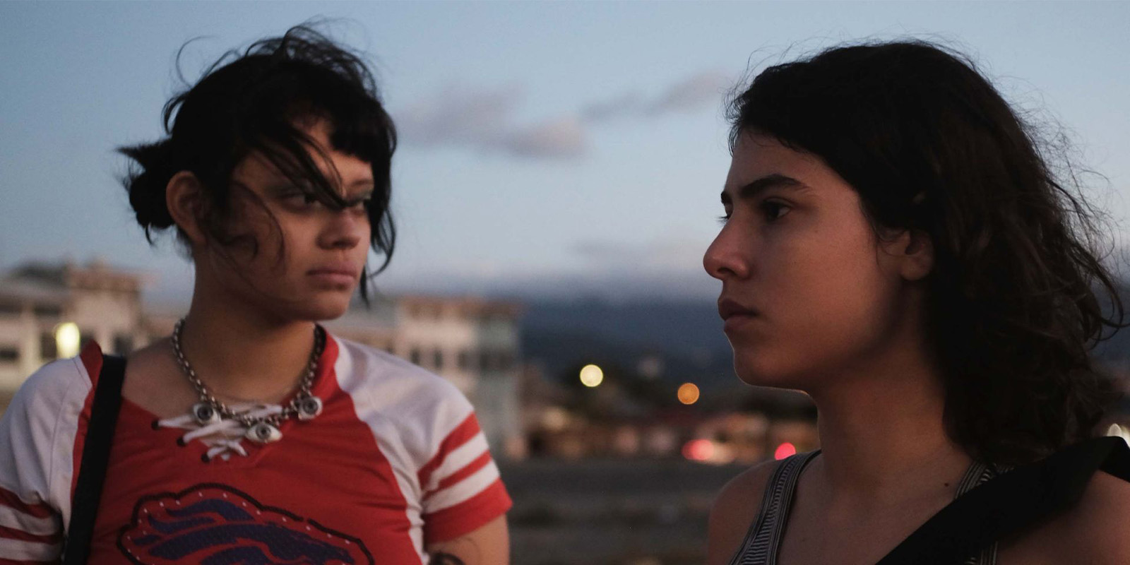 Two people with dark hair stand outdoors at dusk, facing each other. One wears a red shirt with white stripes and a chunky necklace, the other wears a dark vest. Out-of-focus buildings and lights are visible in the background.
