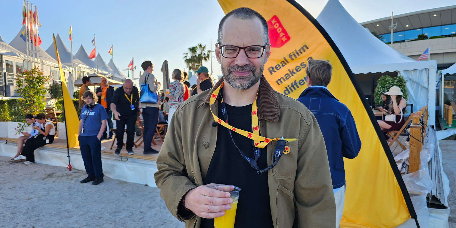 A man holding a cup stands in front of a yellow banner at an outdoor event on a sandy area. Several people are gathered in the background; some are sitting and talking. Flags and tents are visible.