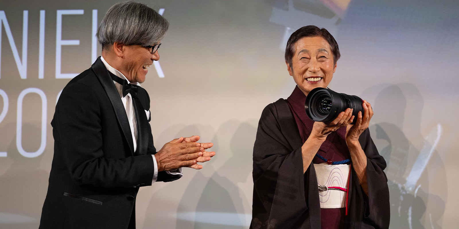 An older woman in traditional Japanese attire smiles whilst holding a large camera lens. A man in a tuxedo stands next to her, applauding. Both are on a stage with a blurred background and large text partially visible.