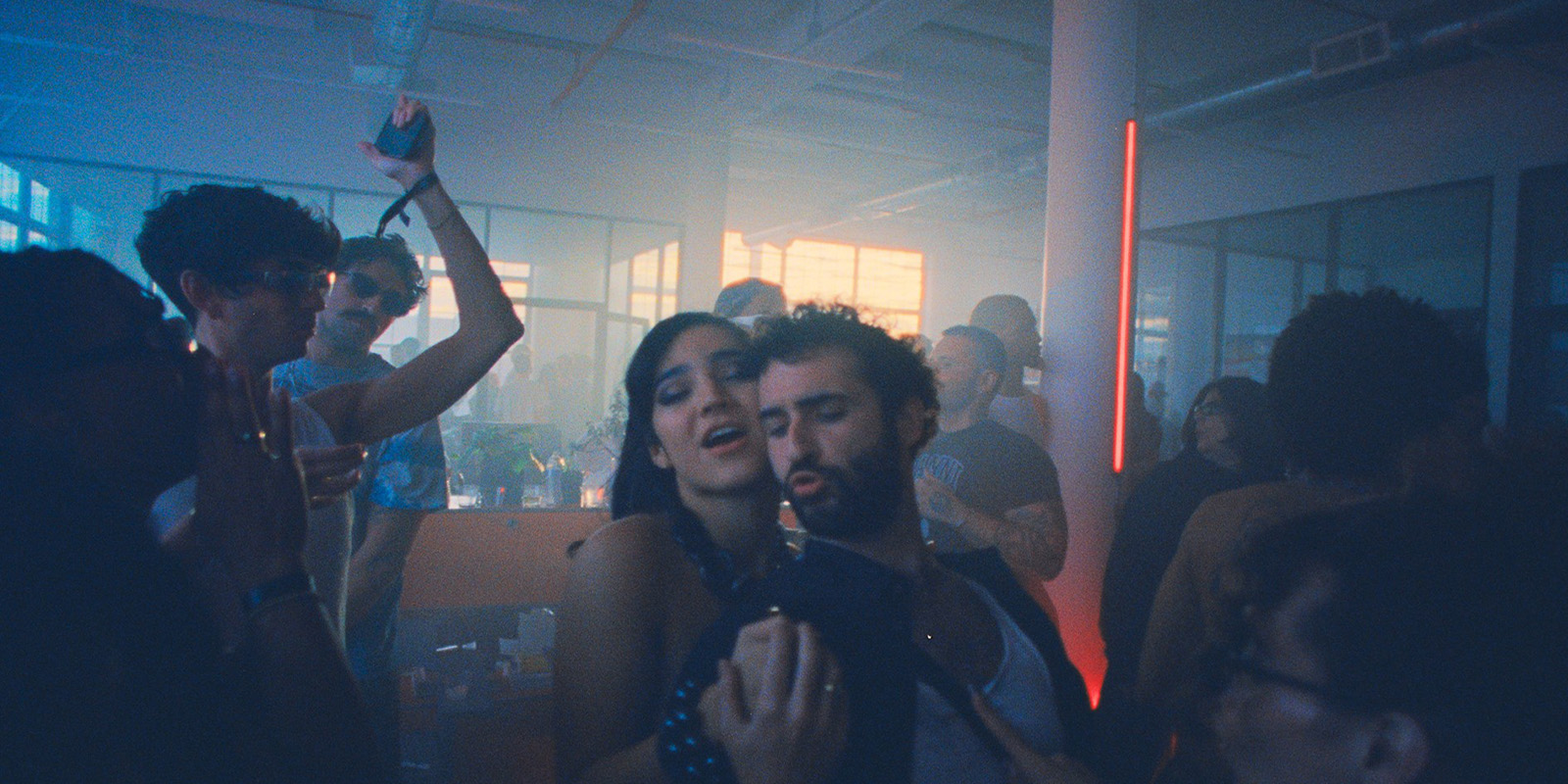 A group of people dance and celebrate at an indoor party with dim lighting. Two people are in the foreground singing and dancing, whilst others enjoy themselves in the background. The room has large windows and neon accent lights.