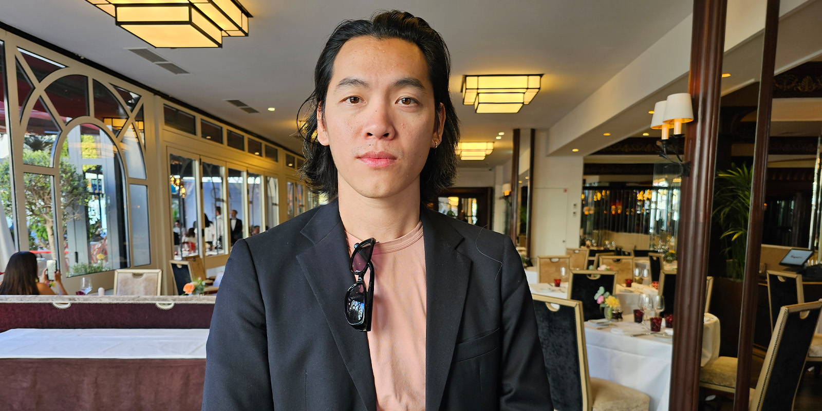 A person with long dark hair, wearing a black blazer over a pink shirt and sunglasses hanging from the collar, stands in a well-lit restaurant with tables laid for dining.