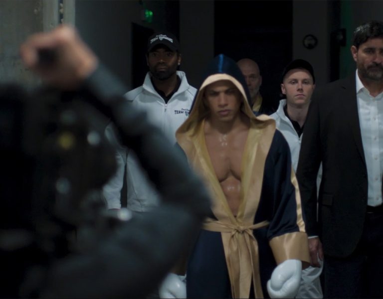 A boxer in a dressing gown and gloves walks down a corridor, flanked by his team and a man in a suit, while a photographer captures the scene.