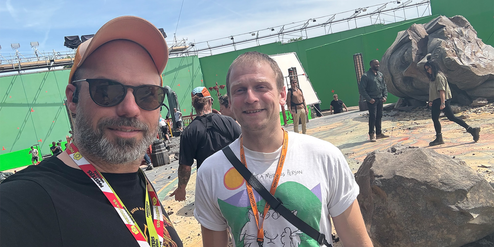 Two people wearing lanyards pose for a selfie on a film set with a green screen, various crew members, equipment, and large artificial rocks in the background.