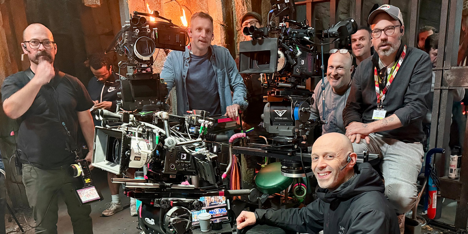 Six people pose behind large film cameras and equipment on a set with barred doors and dim lighting. Some smile at the camera; others look at the equipment. Various gear and tools are visible in the foreground.