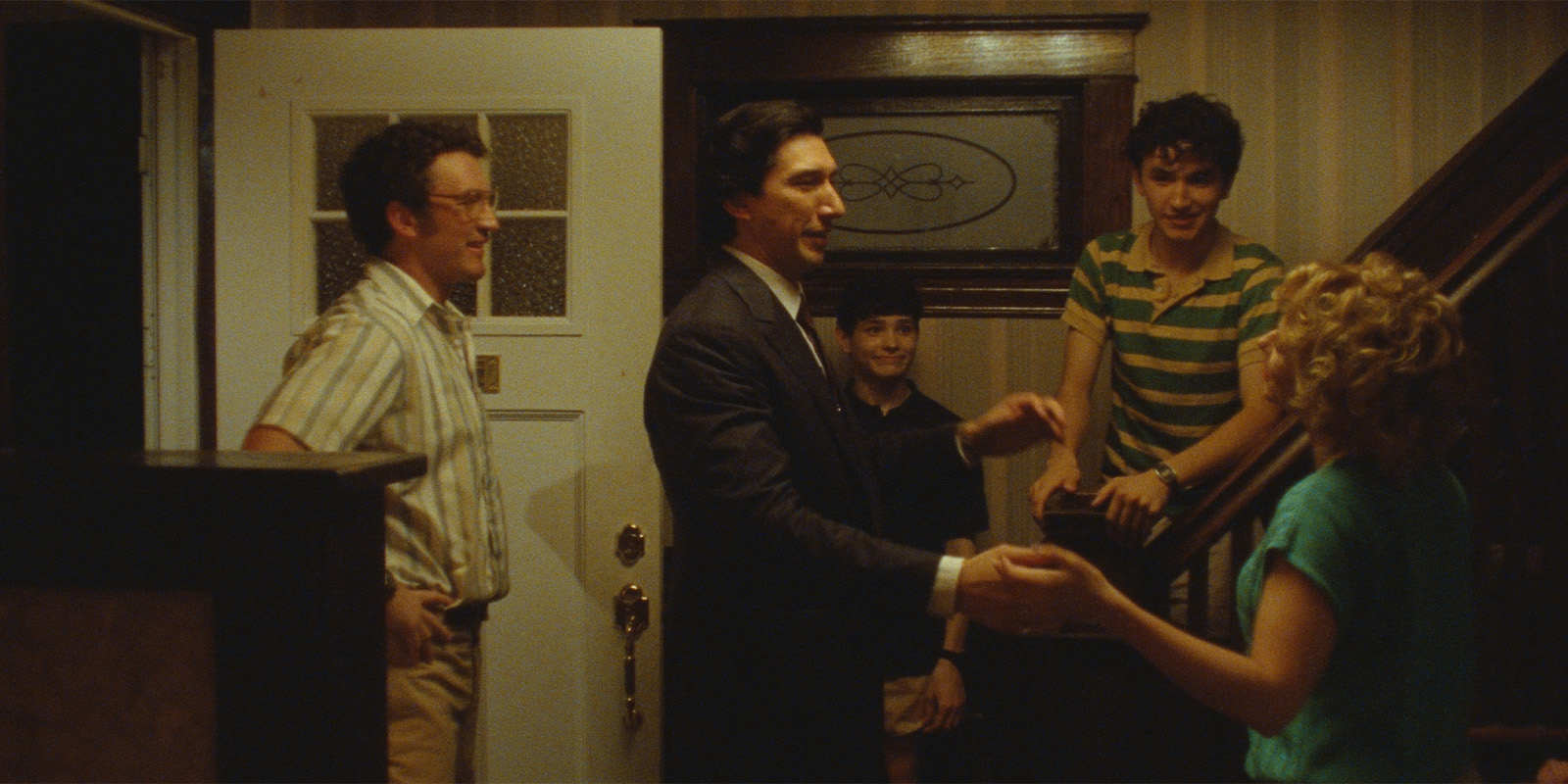Four people greet a woman at the bottom of a staircase inside a house. The man in the centre wears a dark suit, while the others are dressed casually. The front door is open, and the room appears warmly lit.