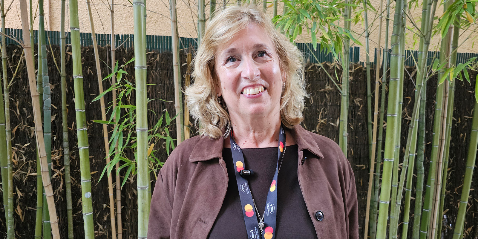 A woman with blonde hair and a brown jacket stands in front of green bamboo plants, smiling at the camera. She wears a lanyard with Mastercard logos.