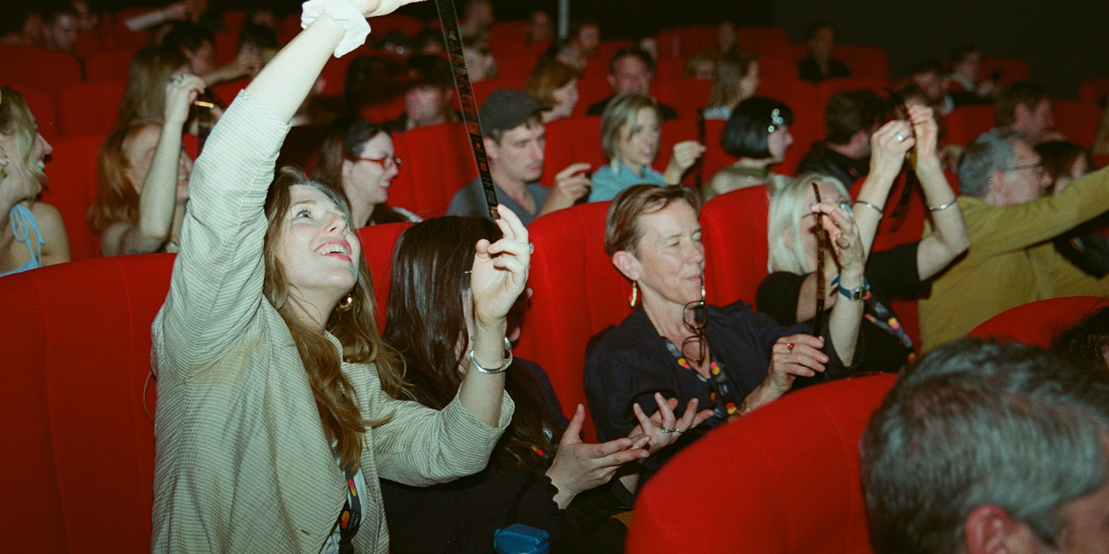 A group of people sit in red theatre seats, holding up strips of film and looking upward. The audience appears engaged and interested, with various individuals focused on examining the film strips.